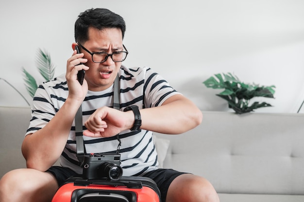 Aziatische reiziger man bril met camera zittend op de bank tijdens het praten op mobiele telefoon en kijken naar horloge in woonkamer Reizen vakantie en vakantie concept