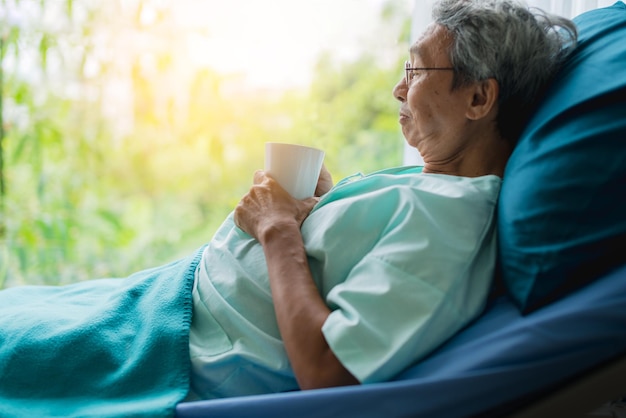 Aziatische senior geduldige man rust uit met hete thee op het concept van gezondheidsideeën in het ziekenhuisbed