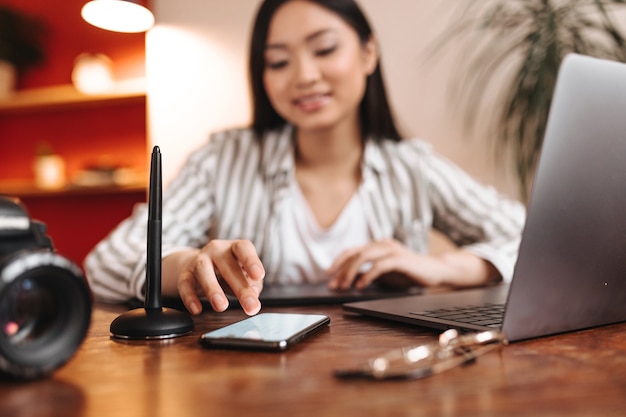 Aziatische vrouw chatten op telefoon met glimlach en poseren op de werkplek met grijze laptop