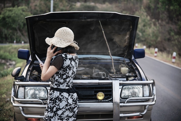 Aziatische vrouw die hersteller of verzekeringspersoneel roepen om een motor van een autoprobleem op een lokale weg te bevestigen