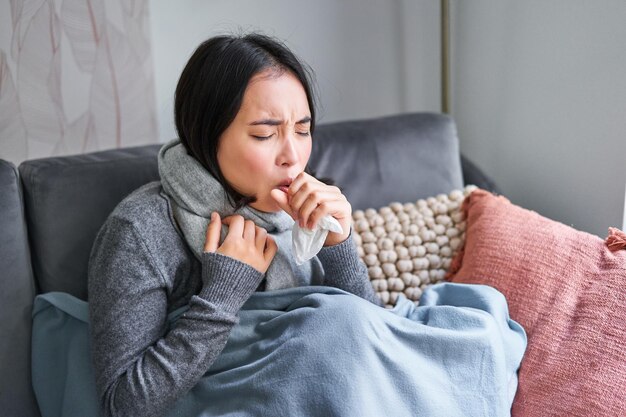 Aziatische vrouw die hoest en zich ziek voelt terwijl ze onder een deken zit met een litteken in een koud appartement, bespaart op warmte