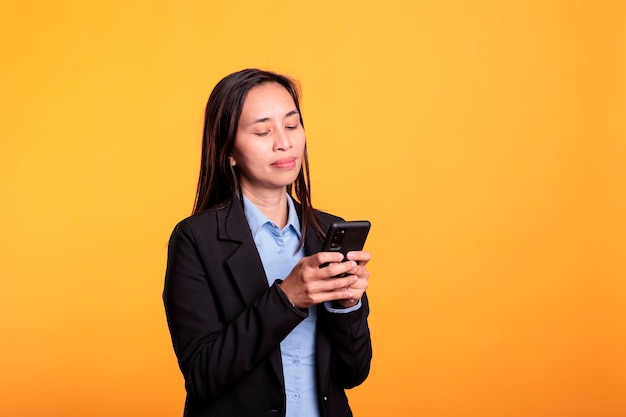 Aziatische vrouw die mobiele telefoon sms-berichten vasthoudt, chatten met externe vriend in studio over gele achtergrond. vrolijk lachende jonge volwassene die geniet van sms-discussie tijdens de opnametijd