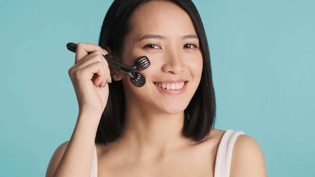 Aziatische vrouw die 's ochtends huidverzorgingsroutine uitvoert en gezicht masseert met een roller die naar de camera lacht over een blauwe achtergrond