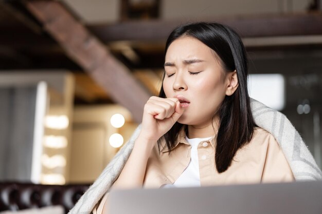 Aziatische vrouw die ziek is, hoest in de vuist, ziek thuis rustend met laptop