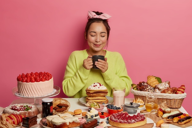 Aziatische vrouw draagt een hoofdband, drinkt thee, omringd met heerlijke desserts, houdt mok vast, houdt de ogen gesloten, geïsoleerd op roze muur. Zoetekauw geniet van een smakelijk ontbijt.