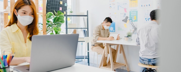 Aziatische vrouw draagt gezichtsmasker voor sociale afstand in een nieuwe normale situatie voor viruspreventie tijdens het gebruik van een laptop en gescheiden door een acryl scheidingswand op kantoor. Leven en werk na het coronavirus.