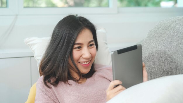 Aziatische vrouw het luisteren muziek en het gebruiken van tablet, het vrouwelijke gebruiken ontspant tijd thuis liggend op huisbank in woonkamer. Gelukkige vrouwelijke het luisteren muziek met hoofdtelefoonsconcept.