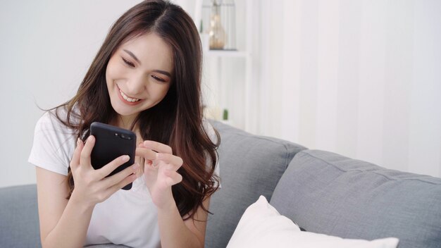 Aziatische vrouw het spelen smartphone terwijl het liggen op huisbank in haar woonkamer.