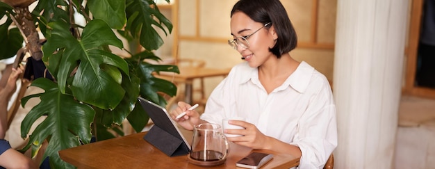 Gratis foto aziatische vrouw in een bril die naar iets kijkt op een digitale tablet, koffie drinkt in een café en glimlachend werkt