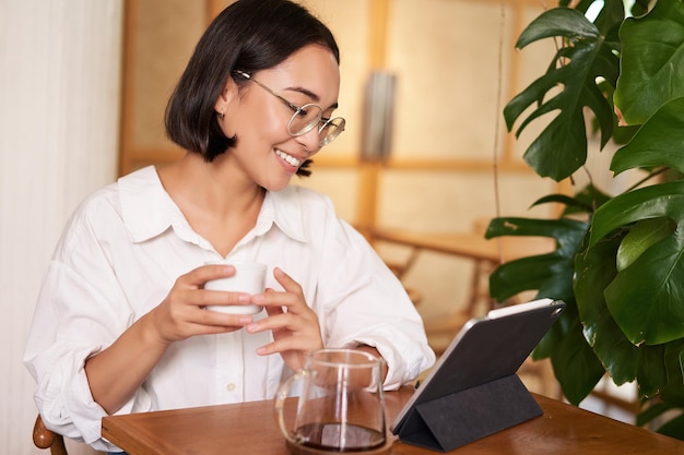 Aziatische vrouw in glazen kijken iets op digitale tablet koffie drinken in een café en glimlachend werken