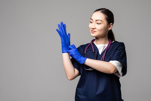 Aziatische vrouw in medisch uniform en gezichtsmasker die beschermende handschoenen draagt die over witte muur worden geïsoleerd