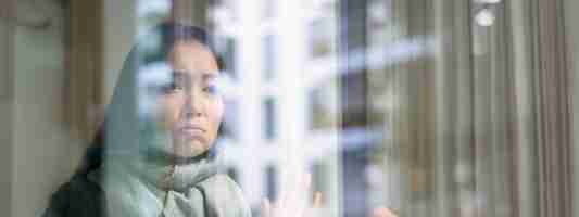 Gratis foto aziatische vrouw voelt zich onwel, blijft thuis, kijkt naar buiten met jaloers, verdrietig gezicht en wil weg