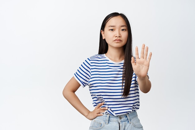 Aziatische vrouw zegt nee. Tienermeisje strekt één hand uit, taboe weigert gebaar, stopbord, fronst en kijkt serieus op wit.