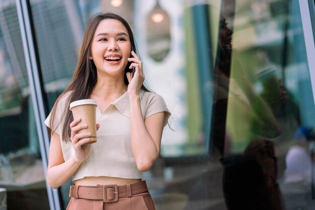 Aziatische vrouwelijke casual ontspannen digitale nomade programmeur kijkt gesprek op smartphone tijdens het wandelen in de stad met koffiekopje glimlachend vrolijk positief gevoel terugschakelen levensstijl