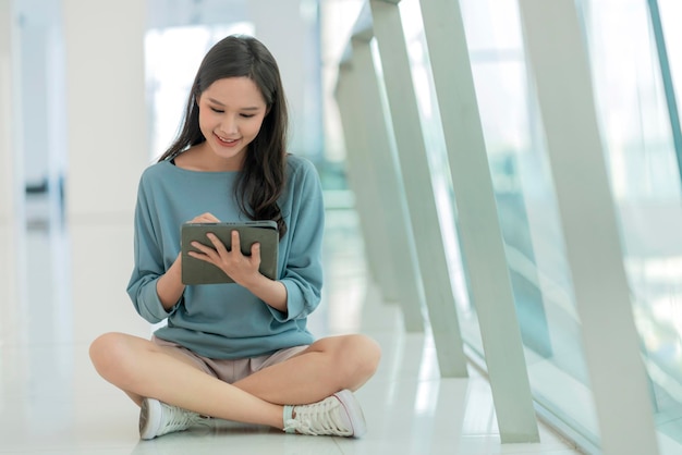 Aziatische vrouwelijke collage jonge campusvrouw zittend op de vloer van het winkelcentrum met behulp van tablet browing surfgegevens online sociale media met vrolijk plezier lachende Aziatische vrouw digitale nomade zit op de vloer casual werk online