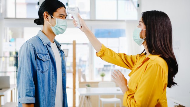 Aziatische vrouwelijke receptioniste die een beschermend gezichtsmasker draagt, gebruikt een infraroodthermometer of een temperatuurpistool op het voorhoofd van de klant voordat hij het kantoor binnengaat. Levensstijl nieuw normaal na coronavirus.