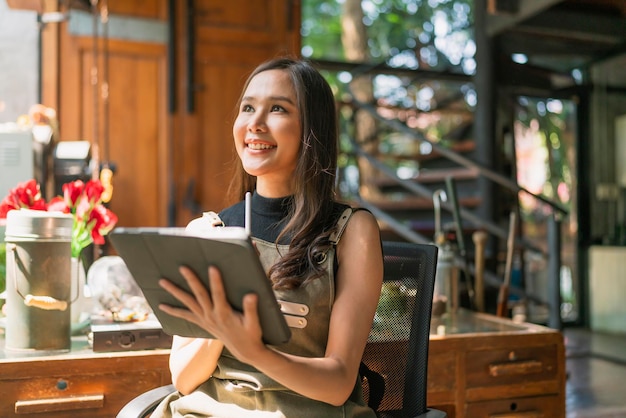 Aziatische vrouwelijke volwassen creatieve ontwerper geluk denkend werk en onderzoek en noteer interessante ideeën via tablet aziatische vrouw die casual werkt ontspannen in haar thuisstudio met raamtuin