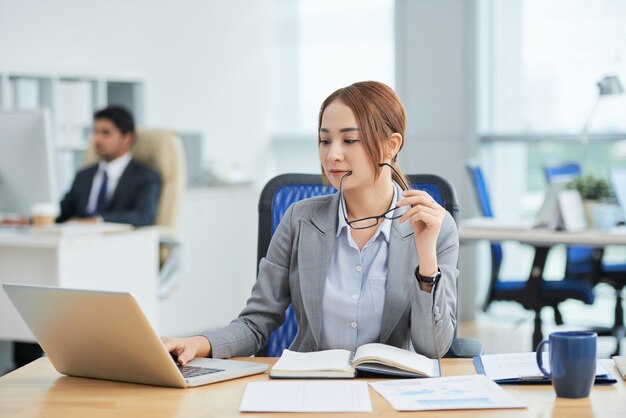 Aziatische vrouwenzitting bij bureau in bureau, holdingsglazen en het werken aan laptop