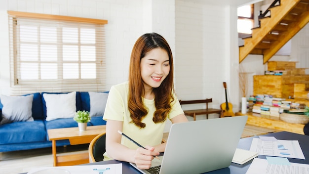 Aziatische zakenvrouw met behulp van laptop praten met collega's over plan in videogesprek terwijl slim werken vanuit huis in de woonkamer. Zelfisolatie, sociale afstand nemen, quarantaine voor coronaviruspreventie.