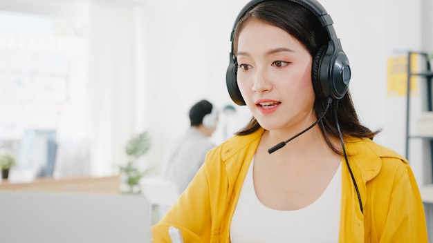 Aziatische zakenvrouw sociale afstand nemen in nieuwe normale situatie voor viruspreventie tijdens het gebruik van laptoppresentatie aan collega's over plan in videogesprek tijdens het werk op kantoor. Leven na het coronavirus.