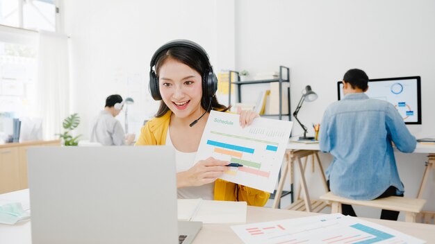 Aziatische zakenvrouw sociale afstand nemen in nieuwe normale situatie voor viruspreventie tijdens het gebruik van laptoppresentatie aan collega's over plan in videogesprek tijdens het werk op kantoor. Leven na het coronavirus.