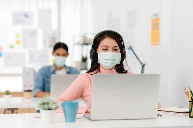 Aziatische zakenvrouw sociale afstand nemen in nieuwe normale situatie voor viruspreventie tijdens het gebruik van laptoppresentatie aan collega's over plan in videogesprek tijdens het werk op kantoor. Leven na het coronavirus.