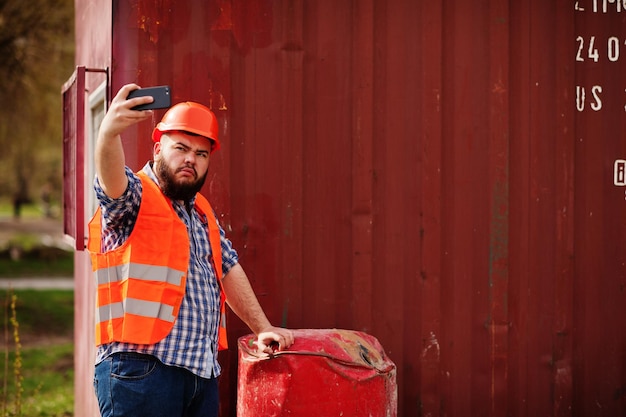 Baardwerker man pak bouwvakker in oranje veiligheidshelm in de buurt van rood vat maakt selfie op telefoon