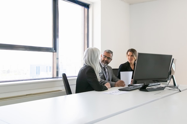 Baas en managers analyseren rapporten en bespreken werk. Team zitten samen op de werkplek met monitoren, papieren en praten. Kopieer ruimte. Zakelijke bijeenkomst concept