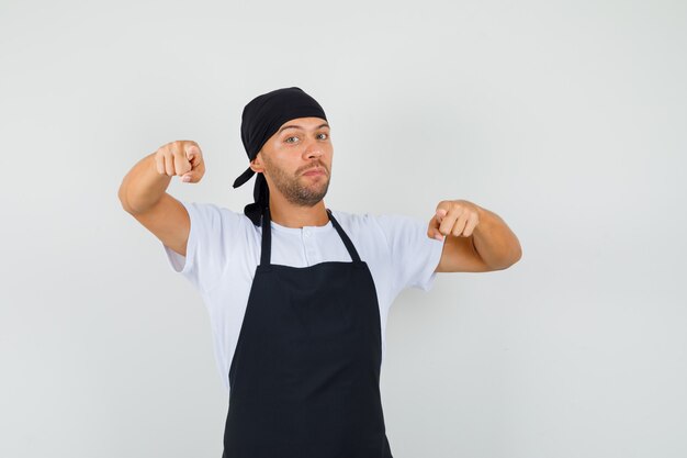 Baker man wijzend op camera in t-shirt, schort en op zoek zelfverzekerd