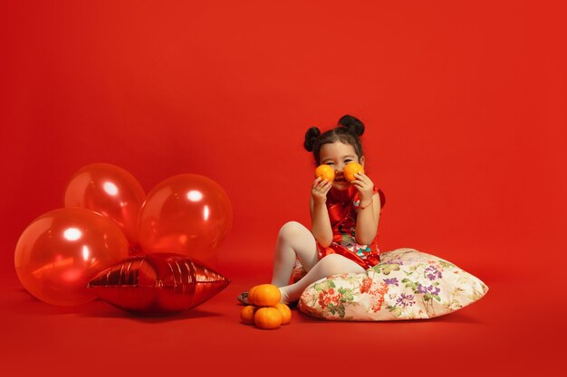 Ballonnen en mandarijnen voor gemoedstoestand. Leuk poseren.​ Aziatisch schattig klein meisje geïsoleerd op rode muur in traditionele kleding. Viering, menselijke emoties, vakantie. Copyspace.