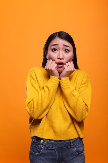 Bang bang jonge volwassen vrouw die mond bedekt met handen in angst op oranje achtergrond. aantrekkelijke brunette aziatische dame was sprakeloos terwijl ze tragisch nieuws op televisie hoorde.