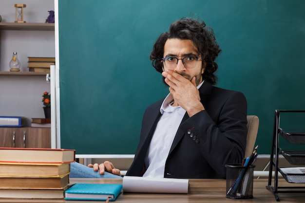 Bang bedekte mond met mannelijke leraar met een bril die aan tafel zit met schoolhulpmiddelen in de klas