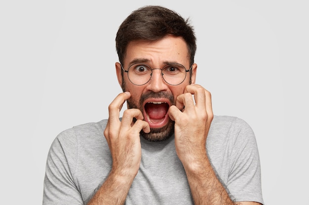 Bang geërgerd mannetje opent mond en fronst zijn wenkbrauwen, houdt de handen op de wangen, kijkt wanhopig, is in paniek, draagt losse grijze kleding, staat alleen tegen een witte muur. Negatieve gevoelens