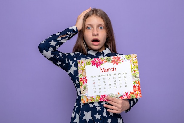 Gratis foto bang greep het hoofd van een mooi klein meisje op de dag van een gelukkige vrouw met een kalender geïsoleerd op een blauwe muur