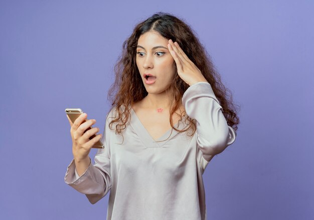 Bang jong mooi meisje houden en kijken naar telefoon en hand zetten voorhoofd geïsoleerd op blauwe muur