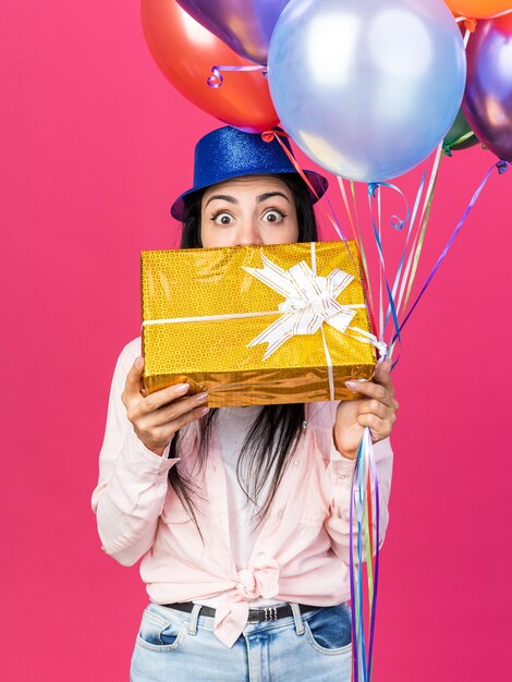 Bang jong mooi meisje met feestmuts met ballonnen en bedekt gezicht met geschenkdoos geïsoleerd op roze muur