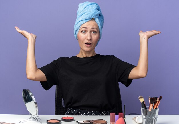 Bang jong mooi meisje zit aan tafel met make-up tools haar afvegen in handdoek spreidende handen geïsoleerd op blauwe achtergrond