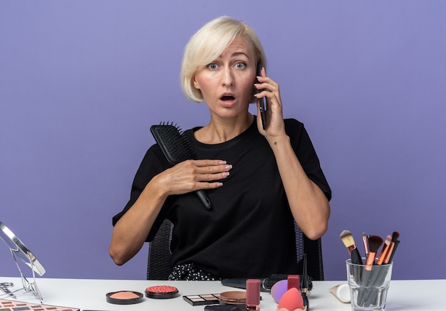 Bang jong mooi meisje zit aan tafel met make-up tools spreekt op telefoon met kam geïsoleerd op blauwe achtergrond