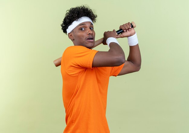 Bang jonge Afro-Amerikaanse sportieve man met hoofdband en polsbandje houden vleermuis op schouder geïsoleerd op groene muur