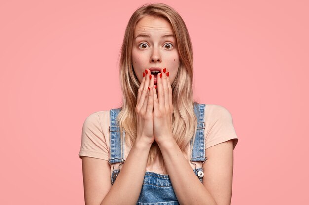 Bang jonge Europese vrouw bedekt de mond met beide handpalmen, is erg emotioneel, reageert op iets geweldigs, heeft rode manicure, gekleed in een casual outfit, geïsoleerd over roze muur