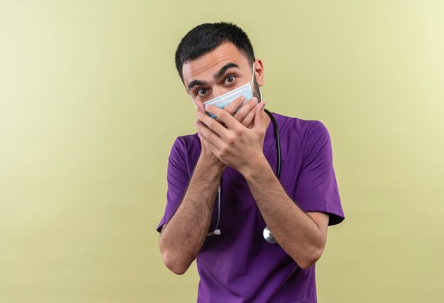 Bang jonge mannelijke arts dragen paarse chirurg kleding en stethoscoop medische masker bedekt mond met handen op geïsoleerde groene achtergrond