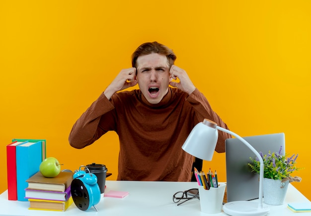 Gratis foto bang jonge student jongen zittend aan een bureau met schoolgereedschap greep hoofd geïsoleerd op gele muur