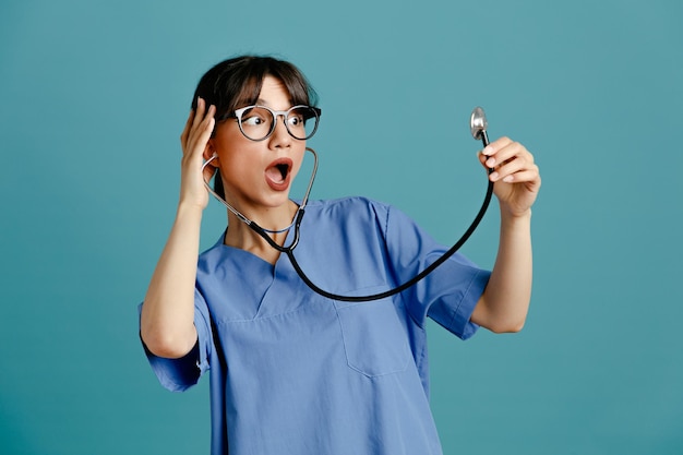 Bang jonge vrouwelijke arts dragen uniform fith stethoscoop geïsoleerd op blauwe achtergrond