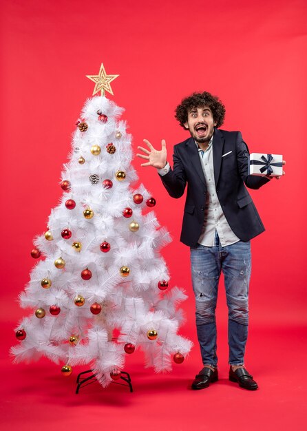 Bang jongeman met zijn geschenk staande in de buurt van versierde witte kerstboom aan de rechterkant van rood