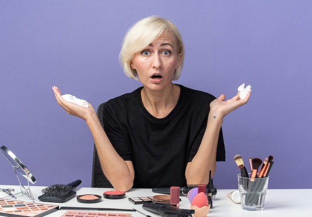 Bang kijkend naar camera zit een mooi meisje aan tafel met make-uptools met haarcrème geïsoleerd op blauwe achtergrond