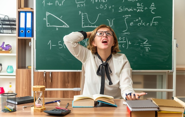 Bang kijkende jonge vrouwelijke leraar die een bril draagt, zit aan tafel met schoolhulpmiddelen die de hand op het hoofd zetten in de klas