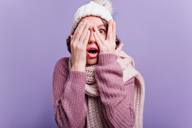 Bang meisje in witte hoed verbergend gezicht met handen. emotionele geschokte vrouw geïsoleerd op paarse lichte muur.