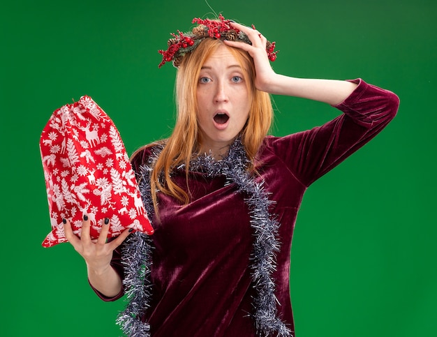 Bang mooi meisje dragen rode jurk met krans en garland op nek met kerst tas hand zetten hoofd geïsoleerd op groene achtergrond