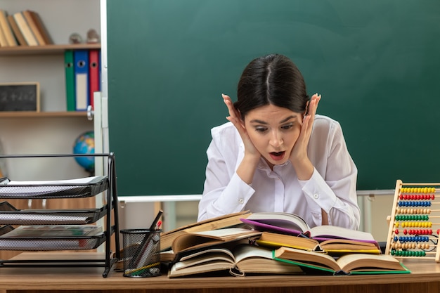 Bang om handen op de wang te leggen, jonge vrouwelijke leraar die een boek leest op tafel die aan tafel zit met schoolhulpmiddelen in de klas