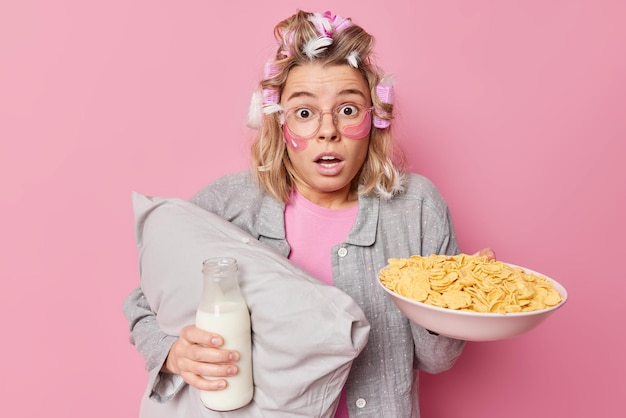 Bang onder de indruk vrouw met haarrollers en veren gekleed in slaappak houdt kussenkom cornflakes en fles melk past hydrogelpleisters toe onder de ogen geïsoleerd op roze achtergrond